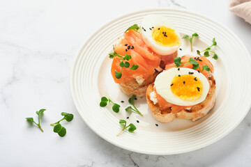 Sandwich with delicious toppings, smoked salmon, eggs, herbs and microgreens radish, black sesame seeds over white plate on white marble table background. Healthy open sandwich superfood. Top view.