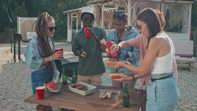 Medium long of company of young diverse people standing around table on beach, making and eating hot dogs on sunny summer day