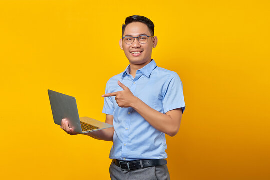 Portrait of cheerful handsome Asian man in glasses pointing finget at laptop,showing something online isolated on yellow background. businessman and entrepreneur concept