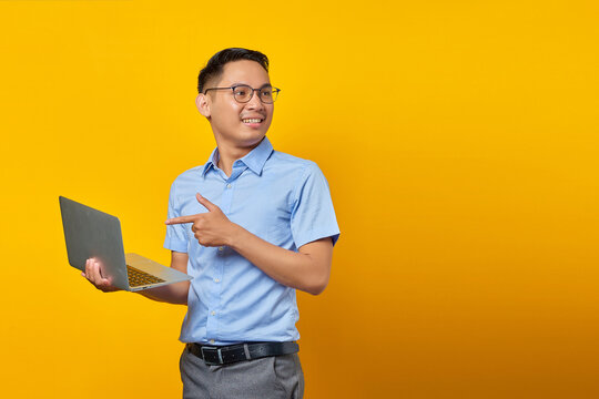 Portrait of cheerful handsome Asian man in glasses pointing finget at laptop,showing something online isolated on yellow background. businessman and entrepreneur concept
