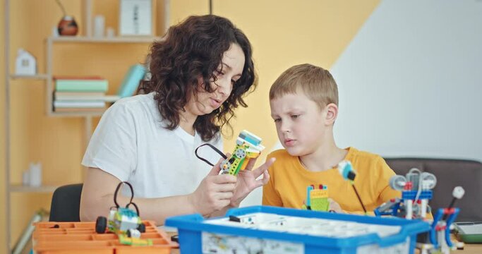 Keeping in mind. Smiling boy sitting with her teacher at the school desk looking at him building a robot out of construction set.