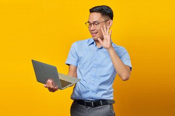 Portrait of cheerful young asian man Asian in glasses using a laptop, doing a video call, gesturing hi to friends isolated on yellow background. businessman and entrepreneur concept