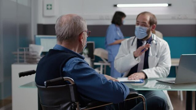 Male patient in wheelchair doing consultation with medic at checkup visit, having conversation about disability and health care. Physician giving advice to impaired senior person with face mask.