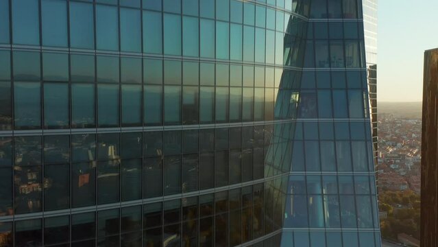 Ascending footage along windows and glass wall of office skyscraper reflecting buildings in town and setting sun. Revealing Spanish flag on rooftop.