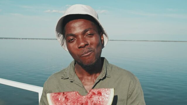 Chest-up of young African American man eating piece of fresh watermelon, standing in foreground of lake on sunny summer day, looking and smiling on camera
