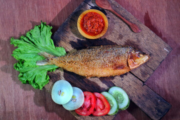 Bandeng Presto Fried Milkfish on the cutting board with salad onion cucumber tomatoes and chili sauce