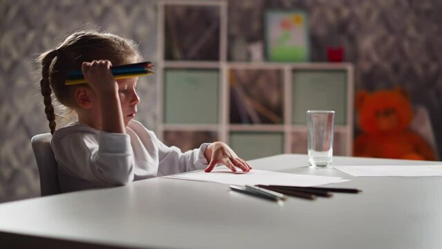 Thoughtful little artist in white shirt turns paper and draws scrabble picture with pencils bunch sitting at desk in children room slow motion