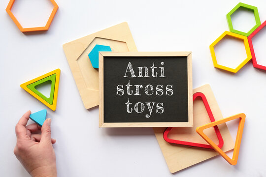 Text Anti stress toys on white blackboard. Nested wood triangles and hexagons in rainbow colors. Turquoise triangle in human hand. Flat lay on white.
