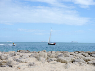 A sailing yacht passing close to the shore with three anchored cruise liners in the distance and a jet ski passing by