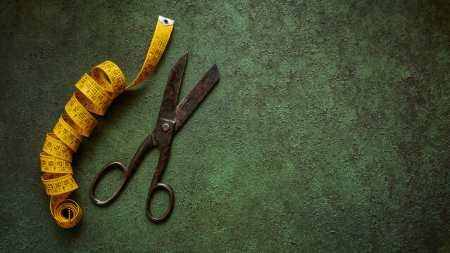 Antique tailor scissors and a tape measure on dark green textured background, flat lay, wide view with copy space