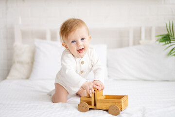 smiling baby 6 months old blond boy is sitting on a large bed in a bright bedroom and playing with a wooden toy car in a cotton bodysuit, the concept of children's goods