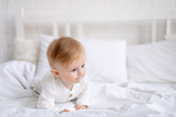 portrait of a 6-month-old blond boy sitting on a large bed in a bright bedroom and smiling in a cotton bodysuit, the concept of children's goods