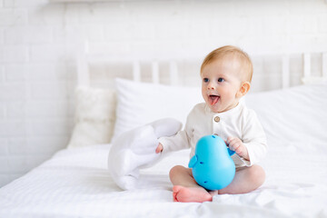 cute baby 6 months old blond boy in a crib in a bright bedroom in a white cotton bodysuit with a blue toy, the concept of children's goods