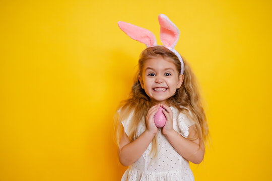 naughty smiling blonde girl with bunny ears holding an easter egg in her hands, on a yellow background studio, space for text, kid celebrate easter