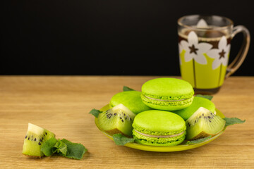 Green cakes macaroons with kiwi slices and mint leaves in saucer on around wooden textured background. A cup of aromatic coffee and dessert. Cooking concept delicious sweet food.