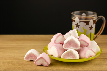 Colored marshmallows sweets in the saucer on around wooden textured background. A cup of aromatic coffee and dessert pink and white in the form of hearts. Cooking concept delicious sweet food.