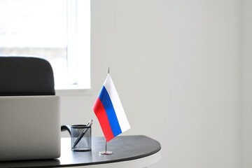 Modern laptop, stationery holder and Russian flag on table in light office