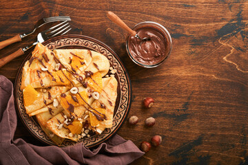 Crepes suzette with oranges and chocolate. Thin crepes with chocolate spread, hazelnuts and orange slices fruit in plate for breakfast on old dark wooden rustic table background. Copy space. Top view