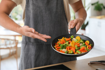 Young man showing frying pan with vegetables in kitchen, closeup