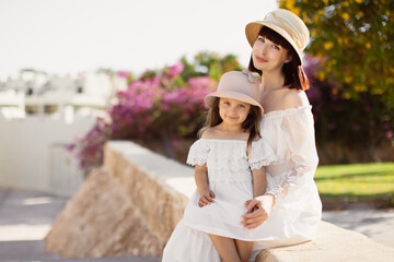 Lovely smiling lady with her daughter posing with straw hat sitting on blur background. Close-up portrait of young mother and her little girl spending time in tropical town in summer morning.