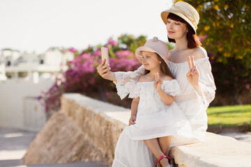 Portrait of happy beautiful caucasian mom and her little cute girl using mobile phone while sitting on the background of tree with flowers and sunny summer landscape, smiling, show a sign of peace