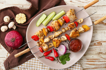 Plate of grilled chicken skewers with vegetables and sauce on white wooden background