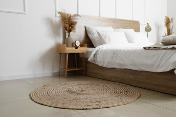 Comfortable bed, wicker rug and nightstand table with dry reeds near white wall
