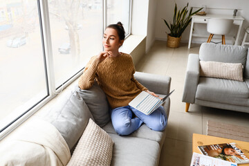 Beautiful young woman with laptop resting on couch at home