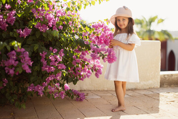 Beautiful little cute girl with long blond hair in white dress and straw hat enjoying pink spring blooming. Little preschool girl in near tree with flowers. Springtime.