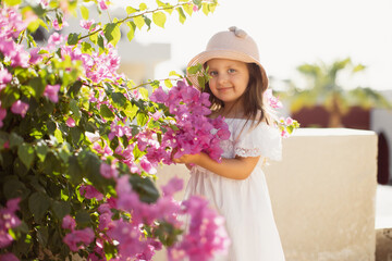 Beautiful little cute girl with long blond hair in white dress and straw hat enjoying pink spring blooming. Little preschool girl in near tree with flowers. Springtime.