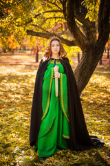 Naklejka premium A beautiful girl in a medieval green dress with gold braid holds a dagger in her hands. Queen in a cloak and in a blio dress in the autumn forest.