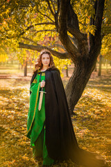 A beautiful girl in a medieval green dress with gold braid holds a dagger in her hands. Queen in a cloak and in a blio dress in the autumn forest.