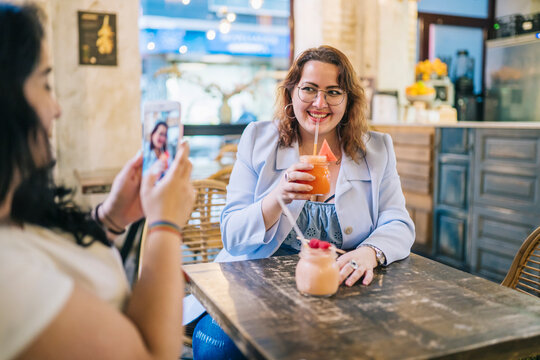 Smiling lady photographing friend drinking juice in bar