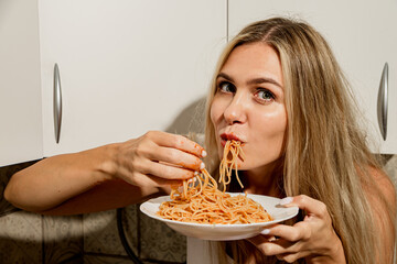 Daring sexy long-legged beauty in a white bodysuit eats spaghetti with tomato paste with her hands. Modern housewife eats pasta in a modern scandinavian kitchen - obrazy, fototapety, plakaty