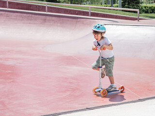 Little boy rides kick scooter in skate park. Special concrete bowl structures in urban park....