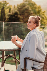 A middle-aged woman sits in a street cafe overlooking the mountains at sunset. She is dressed in a blue jacket and drinks coffee admiring the nature. Travel and vacation concept.