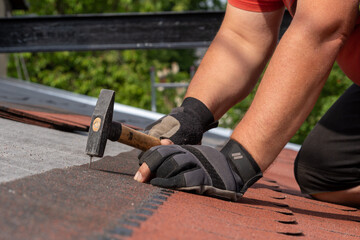 Roofer hands installing bitumen roof shingles using hammer in nails. Installation of waterproofing...