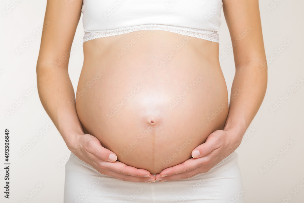 Canvas Prints Young adult pregnant woman in white clothes touching big naked belly with hands. Showing shape. Pregnancy concept. Expectation time. Closeup. Isolated on light gray wall background.