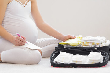 Young adult pregnant woman with big belly sitting on carpet, packing and baby clothes and diapers...