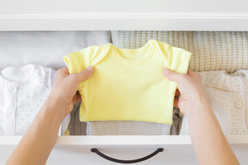 Young adult woman hands holding yellow bodysuit and sorting clothes for baby in white drawer box....