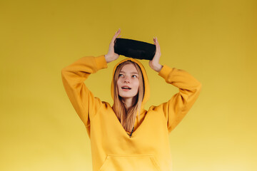 Funny young girl looking, waving hand, holding portable wireless bluetooth music speaker, isolated on yellow background. Girl in a yellow hoodie