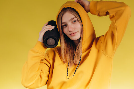 Funny young girl looking, waving hand, holding portable wireless bluetooth music speaker, isolated on yellow background. Girl in a yellow hoodie