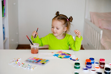 A nice girl is sitting at a table and drawing a picture on the theme of the autism symbol. Heart of puzzles in watercolor on paper. The child learns to draw and color, hold the paintbrush correctly. 