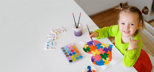Concept of support for children with autism syndrome. Little girl draws a drawing with a picture of a heart from a puzzle with watercolor paints. Painting lessons. Baby at the table holds a paintbrush