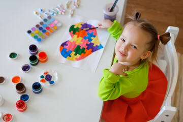 Kid is happy with his drawing as a sign of support for children with autism. Bright multicolored Heart from puzzles as a gift to friend. Art classes with kids. Girl looks at the camera smiling sweetly