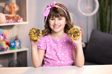 Portrait of sweet girl choosing between two donuts in home room. Cheerful school girl playing with cakes indoors. Funny teenager girl having fun with colorful donuts at modern home