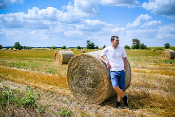 Naklejka na ściany i meble Happy farmer is standing beside bales of hay. He is examining straw after successful harvesting. Strong handsome man portrait on agricultural field