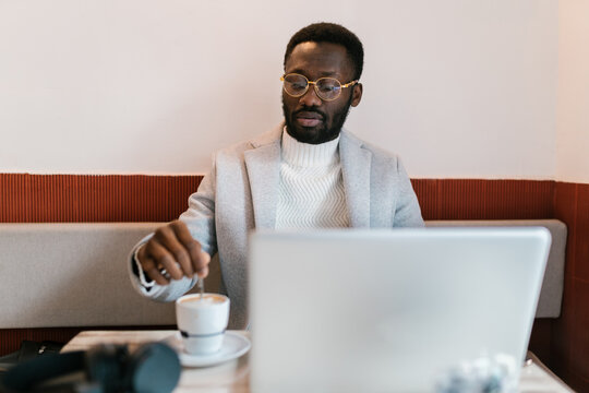 Black businessman stirring coffee near laptop in cafe