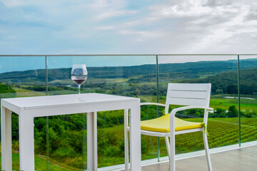 A table with a glass of red wine and a chair with a cushion on an empty balcony with glass railings. White furniture on the terrace overlooking the vineyard.