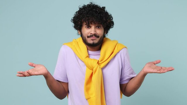 Fun confused shy shamed young bearded Indian man 20s wears violet t-shirt look camera spreading hands say oops ouch oh omg i am so sorry isolated on plain pastel light blue background studio portrait